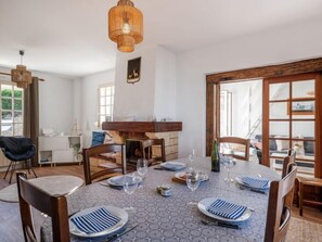 A dining area with a round table set for six, surrounded by wooden chairs. A fireplace is visible in the background, and there is a large window to the right.