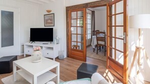 A room with a TV on a white stand, a white table, a black ottoman, and a wooden door leading to another room. - Kergroix by Interhome (Quiberon)