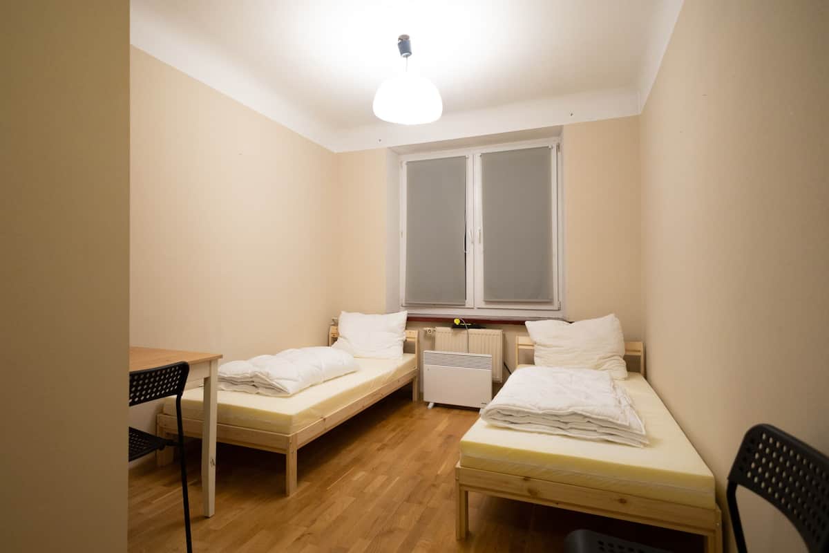 Una habitación pequeña y bien iluminada con dos camas individuales, una mesa de madera, una silla y un radiador.