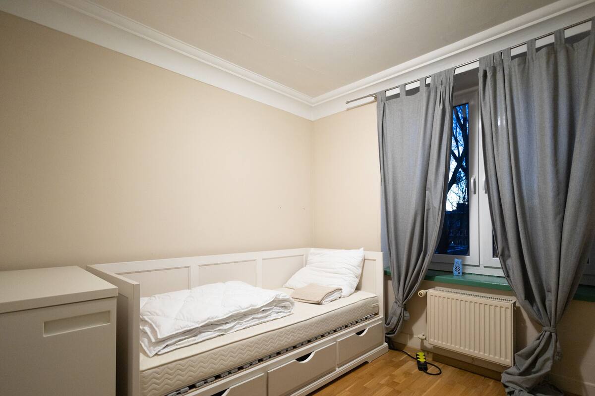 Una habitación pequeña con cama individual, ventana y cortinas, un radiador y un armario.