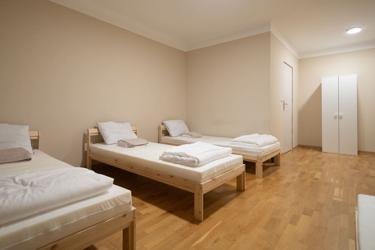 Una habitación con tres camas de madera, cada una con ropa blanca y almohadas. Hay un armario blanco y una puerta a la derecha.