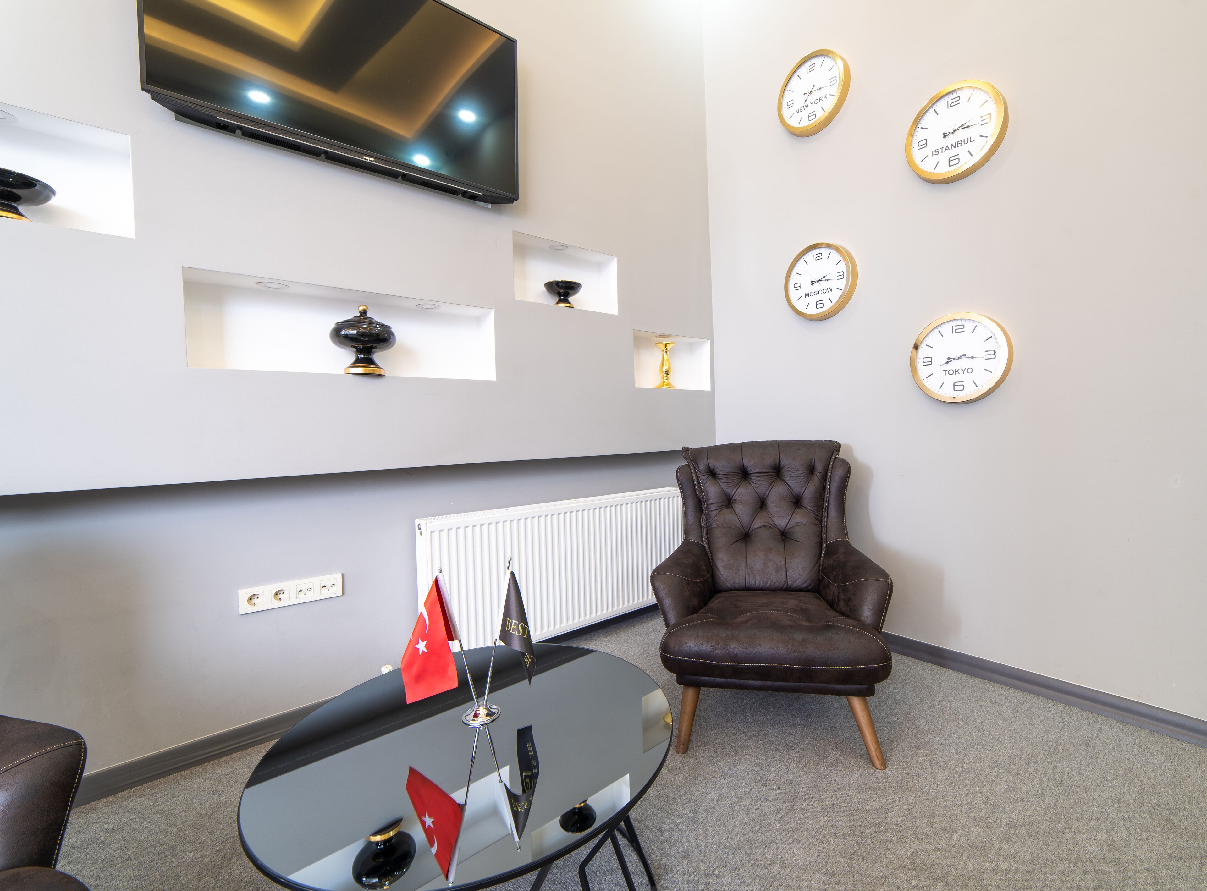Un espacio de oficina moderno con un sillón de cuero, una mesa de vidrio con banderas, un televisor montado y relojes que muestran la hora de diferentes ciudades.