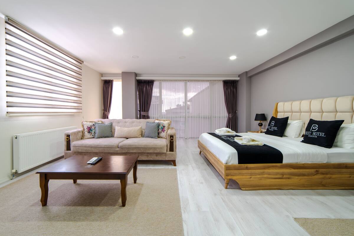 A modern hotel room with a sofa, a wooden bed, a coffee table, and a window with blinds.