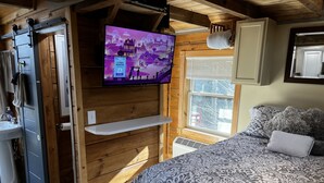 Ein gemütliches Zimmer mit einem Bett, einem Wand-Fernseher, einem Fenster mit Jalousien, einer Holzbalkendecke und einem Regal.