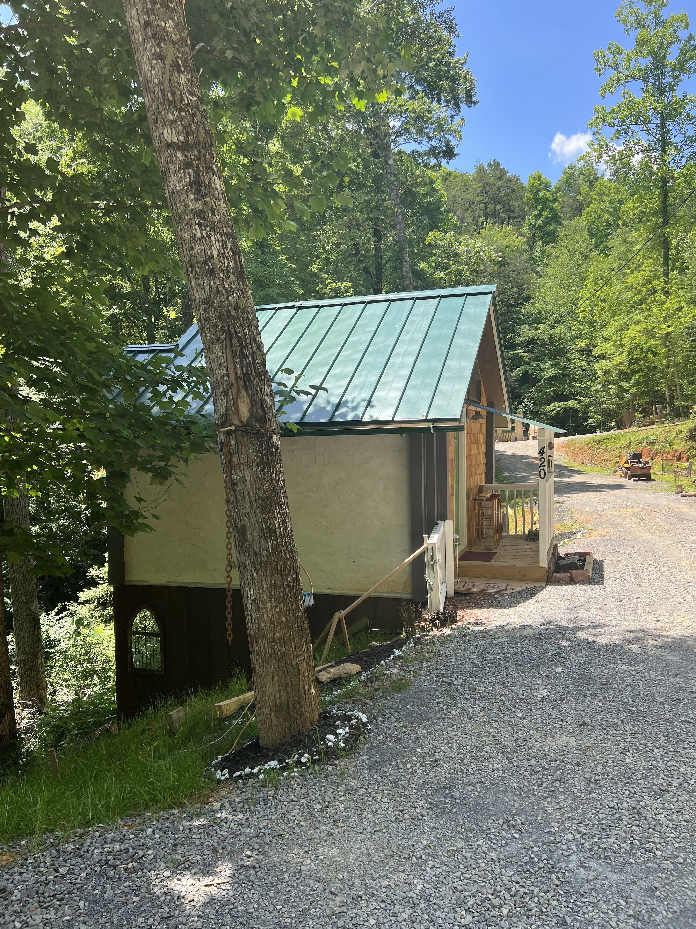 A small, single-story building with a green metal roof, situated on a gravel path surrounded by trees.