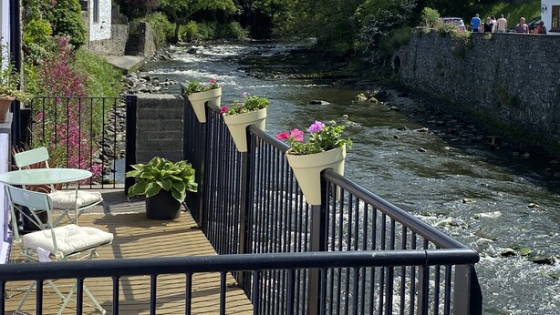 Ein Deck am Flussufer mit Außenbestuhlung, einer Steinmauer und Blick auf einen Fluss.