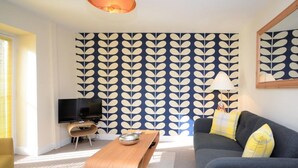 Ein modernes Wohnzimmer mit strukturiertem Wandpapier, einem Flachbildfernseher, einem Holztisch und einem grauen Sofa mit gelben Kissen.