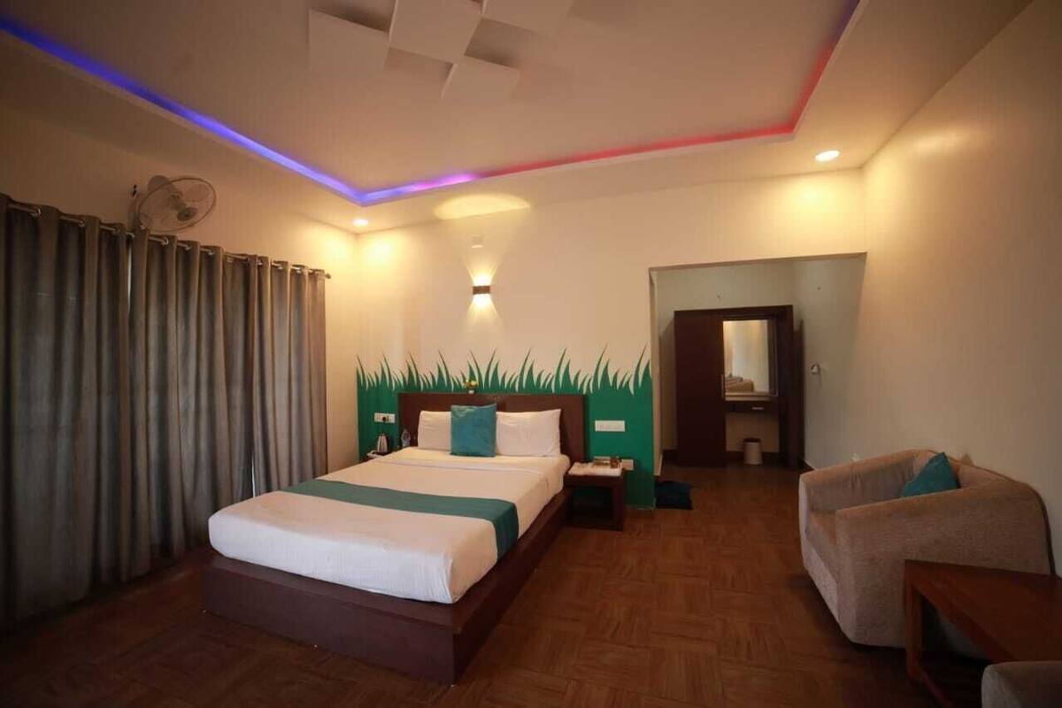 A hotel room with a bed, a chair, a small table, a nightstand, a mirror, and a wall-mounted fan.