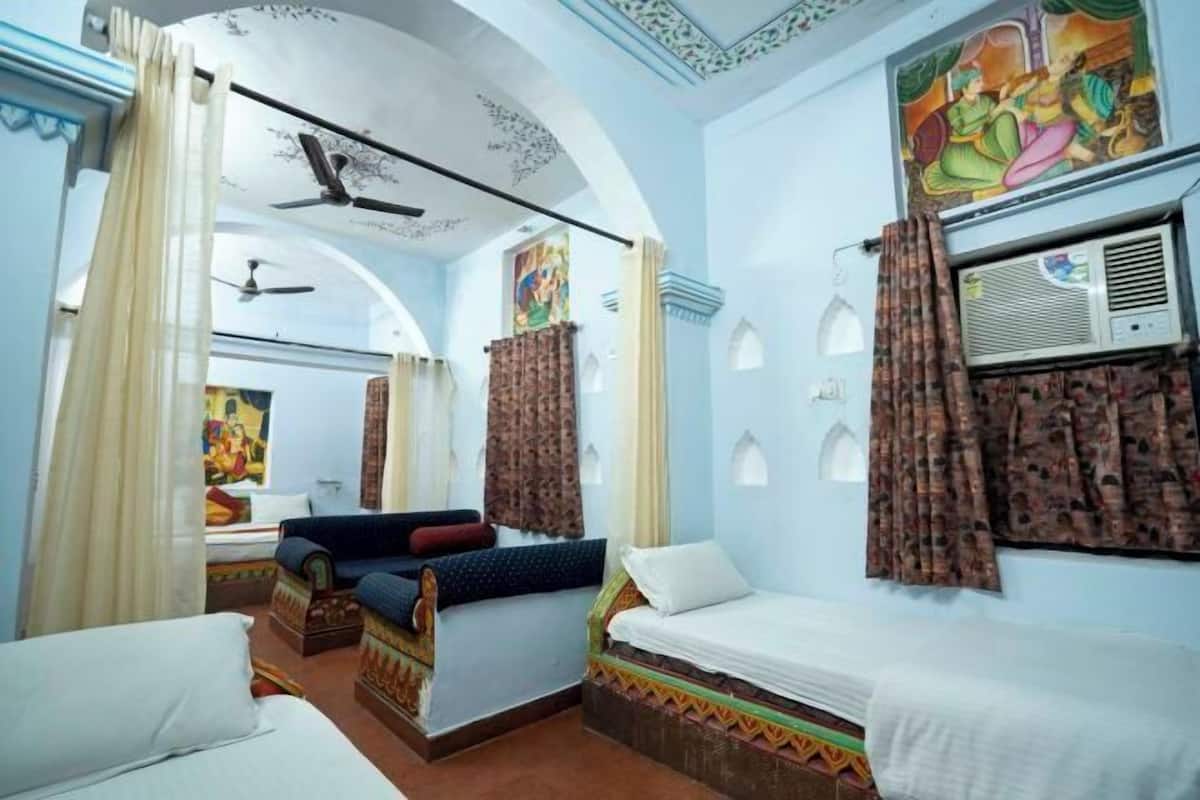 A room with two beds, a ceiling fan, a window with curtains, and a wall with a colorful painting.