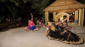 A group of women performing a traditional dance in an outdoor setting with a thatched-roof pavilion.