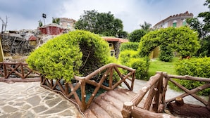 A rustic wooden bridge over a stone-paved area with lush greenery and a stone building in the background.