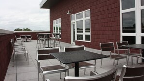 An outdoor patio area with multiple tables and chairs, a red building with large windows, and a cloudy sky.