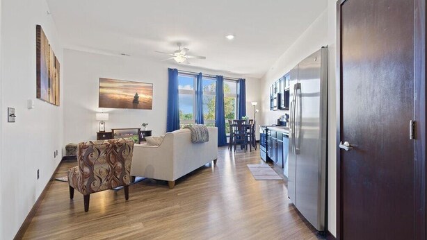 A spacious living area with a sofa, a patterned armchair, and a dining table. A kitchen with a refrigerator and cabinets is visible in the background.