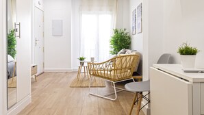 A modern, well-lit room with a wooden floor, a wicker chair, a small table with a plant, and a desk with a chair.