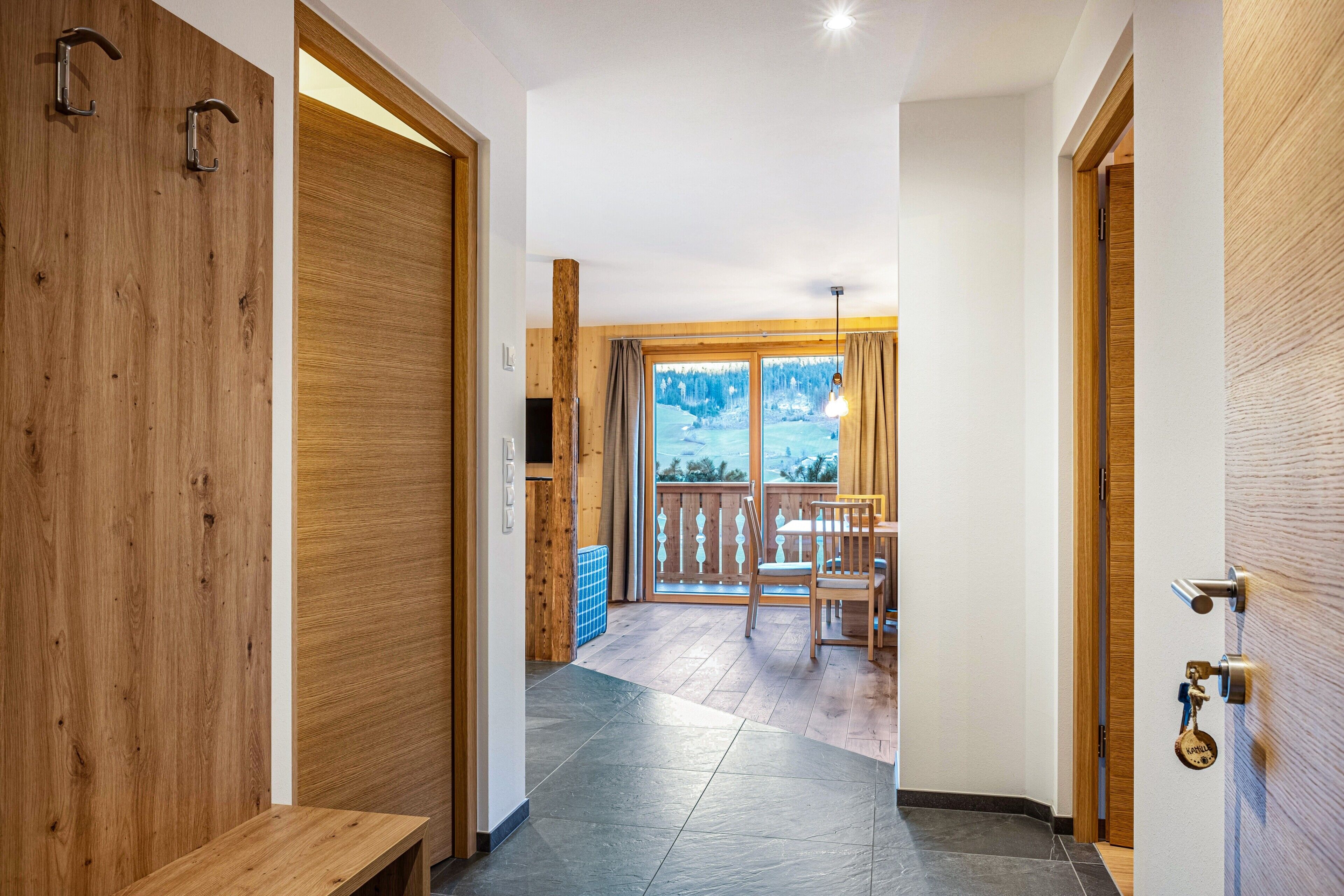 A hallway with wooden doors, a bench, a dining area with chairs and a table, and a view of a snowy landscape through large windows.