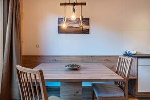 A dining area with a wooden table, chairs, a bowl of herbs, and a painting of a sunset on the wall.