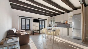 A modern kitchen with white cabinets, a built-in oven, and a refrigerator. There is a dining area with a table and chairs, and a sofa in the living area.