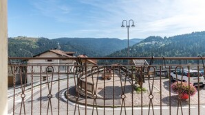 Varanda com grade de ferro forjado, vista para área pavimentada com rotatória, um edifício e paisagem montanhosa.