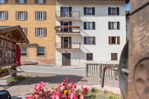Un edificio de varios pisos con balcones, una calle en primer plano y una fuente.