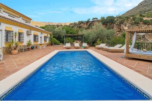 Piscina retangular com borda em azulejo, rodeada por espreguiçadeiras e um pérgola.