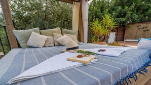 Una cama con una manta azul y blanca a rayas, un libro, gafas de sol y una planta sobre una plataforma de madera.