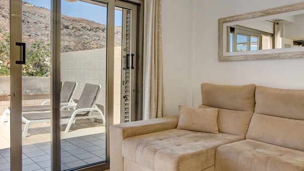 Ein Zimmer mit einer beigefarbenen Couch, einer Schiebetür zu einem Balkon mit zwei weißen Liegestühlen und einem Spiegel an der Wand.