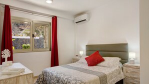 Ein Schlafzimmer mit Bett, Nachttischen, einem Schreibtisch, einem Fenster mit Vorhängen und einer Klimaanlage.