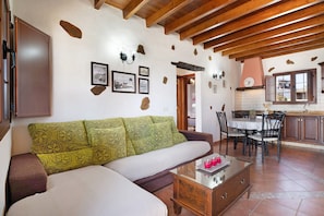 A living room with a sofa, a dining table with chairs, a kitchen area, and a wooden ceiling.