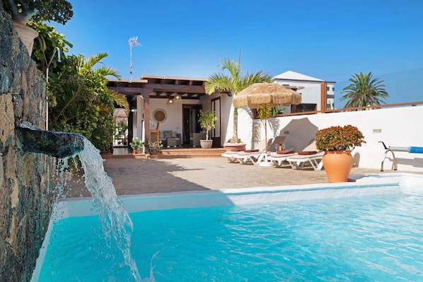 A pool with a waterfall feature, lounge chairs, and a building with a thatched umbrella.