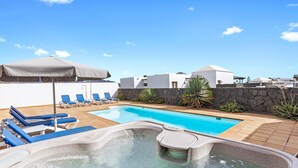 En swimmingpool med solstolar och ett bubbelbad, omgiven av vita byggnader och en stenmur.