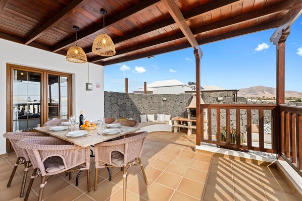 Un patio techado con mesa de comedor, sillas de mimbre, pared de piedra y vista al paisaje.