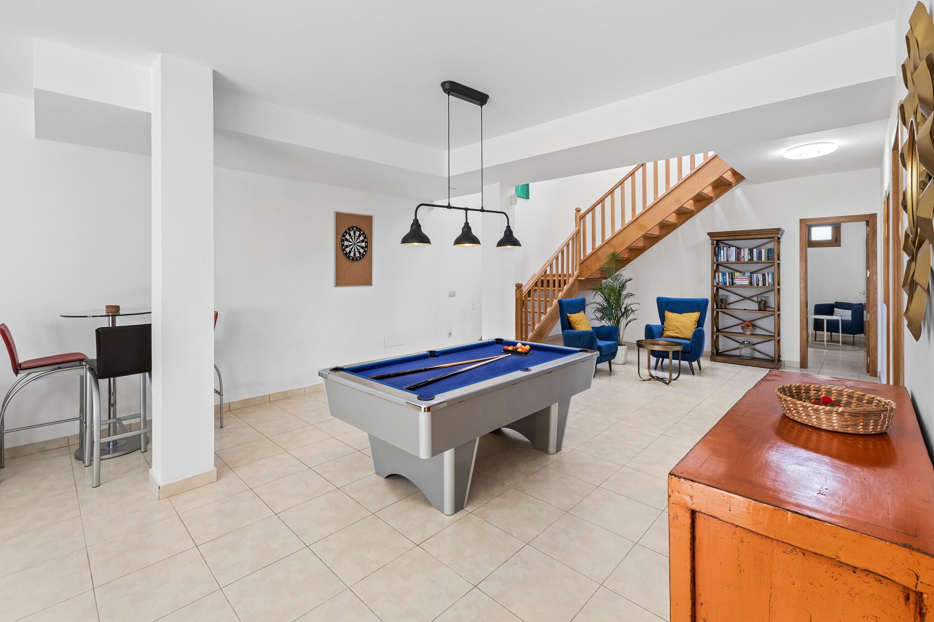 A pool table in a room with a staircase, bar stools, and a bookshelf.
