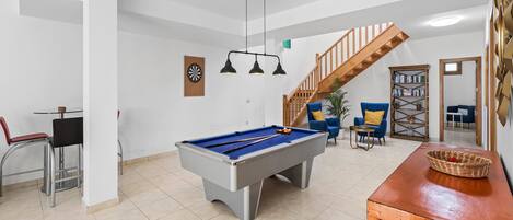 A pool table in a room with a staircase, bar stools, and a bookshelf.