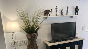 Una sala de estar con televisor, una planta decorativa, una lámpara y una estantería con figuritas.