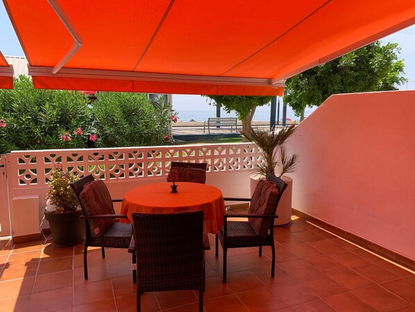 Een terras buiten met een ronde tafel, vier stoelen, een bloempot en uitzicht op zee.