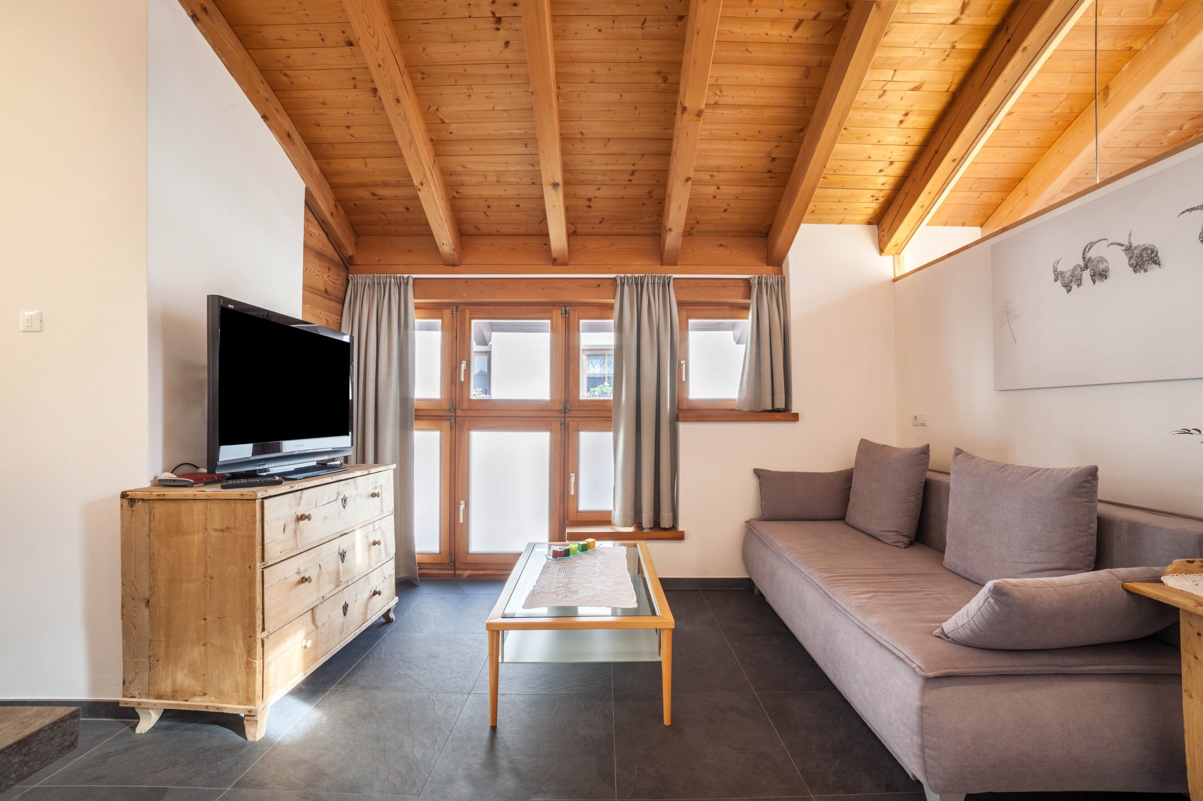 A living room with a wooden ceiling, a television on a wooden cabinet, a sofa, and a window with curtains.