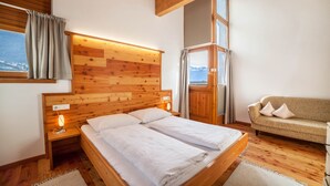 Ein Schlafzimmer mit Holz-Kopfteil, einem Bett mit weißen Leinentüchern, einem Nachttisch mit Lampe, einem Sofa und Blick auf Berge durch ein Fenster.