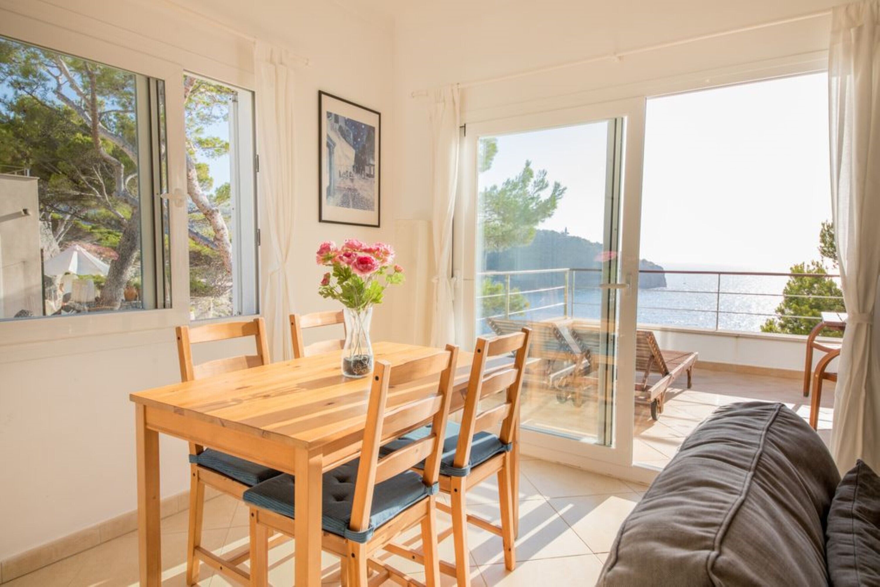 Un comedor con una mesa y sillas de madera, un jarrón con flores y vistas al mar a través de ventanas grandes.