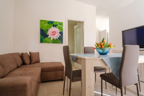 A living room with a sofa, a dining table with chairs, a TV, and a painting of flowers.