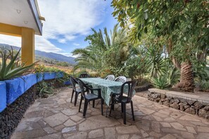 An outdoor patio with a table and chairs, surrounded by plants and a view of mountains.