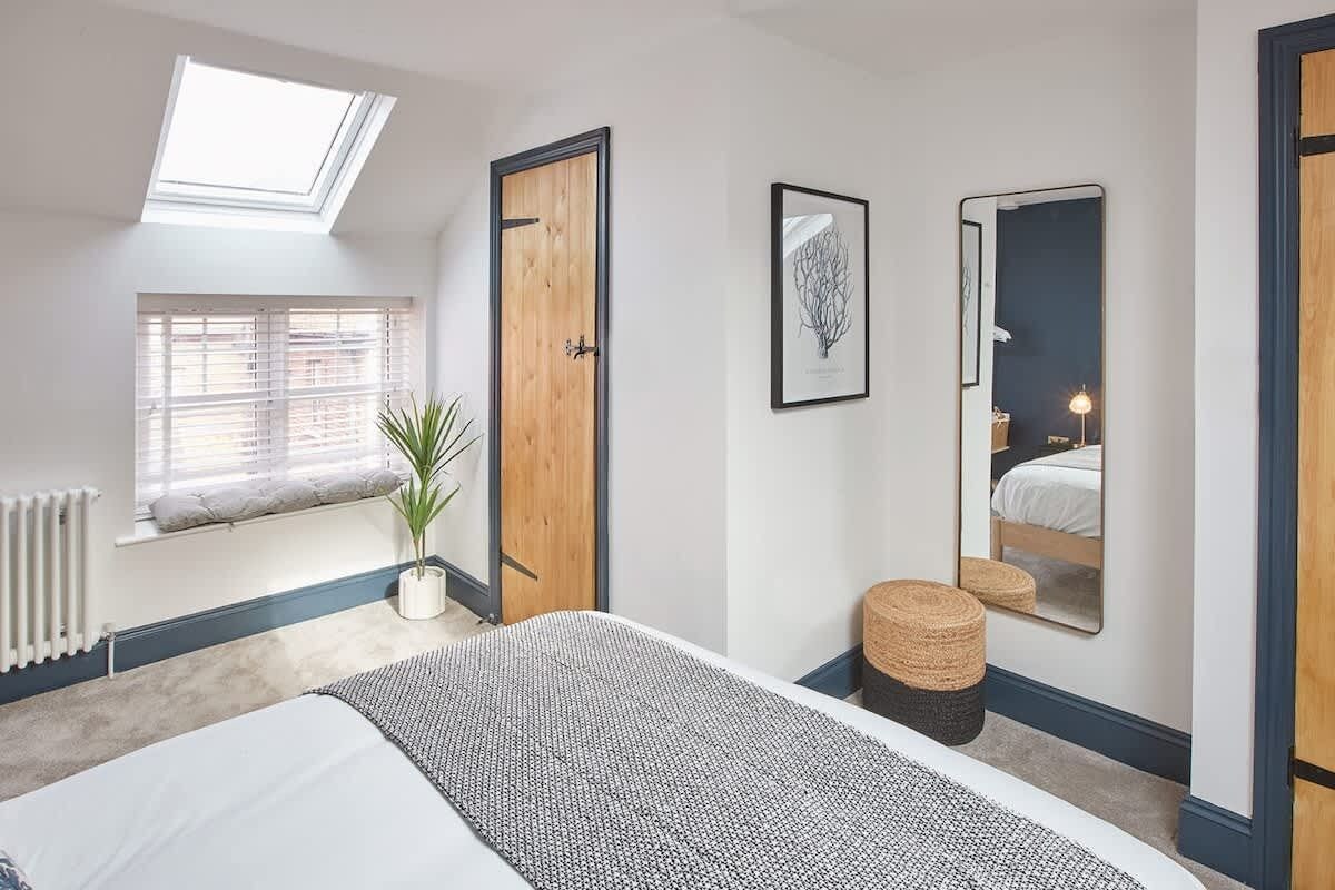 A bedroom with a skylight, a bed, a window with blinds, a plant, a mirror, and a bedside table.
