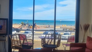 A room with a large window overlooking a beach, a few chairs, and a small table.