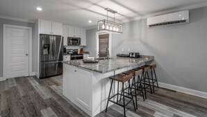 Una cocina moderna con armarios blancos, un refrigerador de acero inoxidable, un microondas y una encimera de granito. Hay tres taburetes de bar y una lámpara colgante.