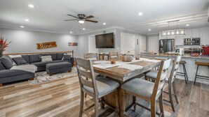 Una sala moderna con un sofá seccional gris, una mesa de comedor de madera con seis sillas, una zona de cocina con electrodomésticos de acero inoxidable y un ventilador de techo.