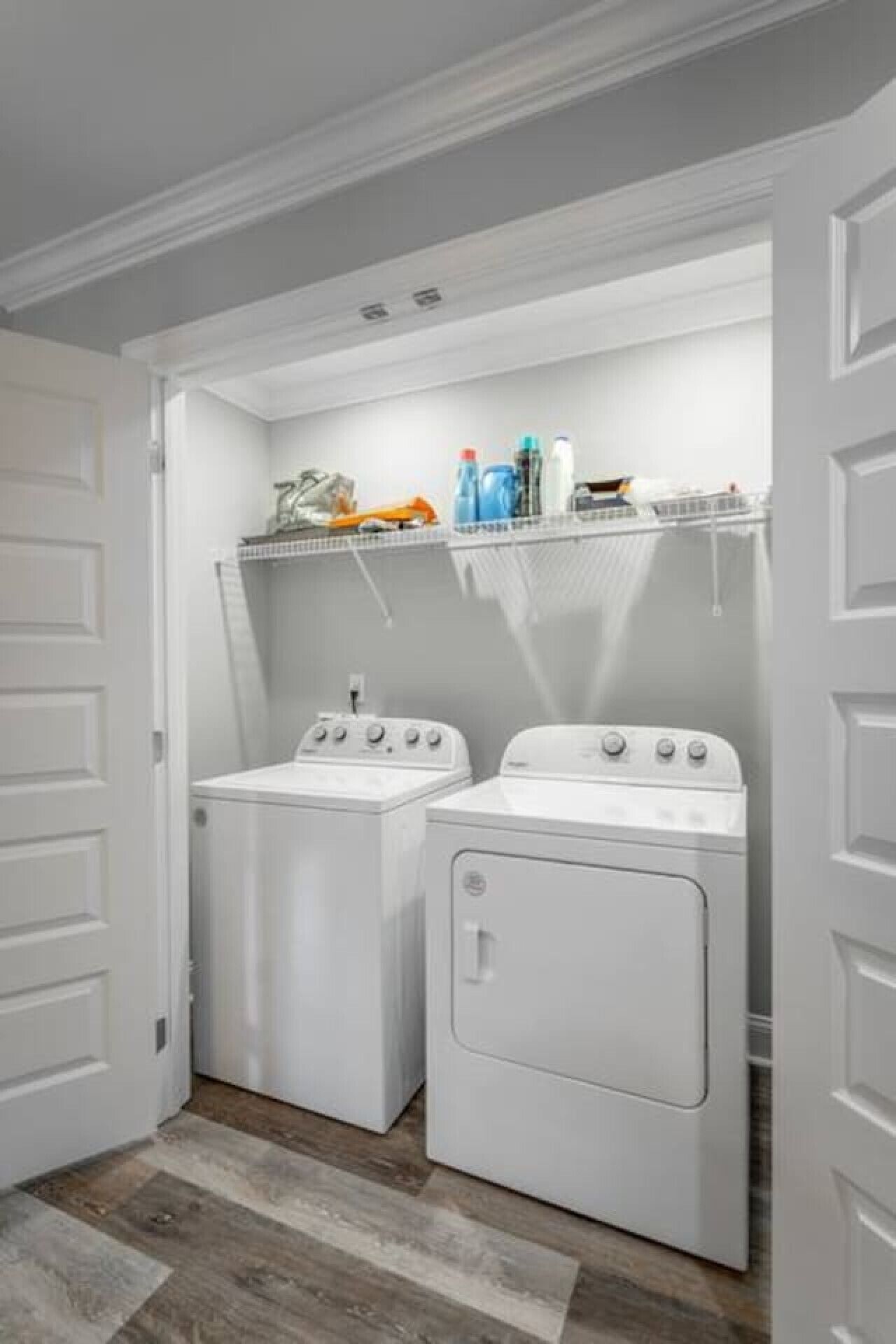 A laundry room with two front-loading washers and dryers, a shelf above with cleaning supplies, and a wooden floor.