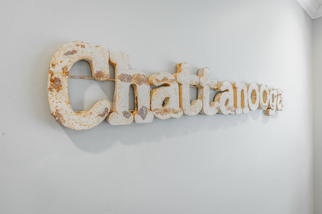 A rustic, weathered metal sign spelling "Chattanooga" mounted on a plain white wall.