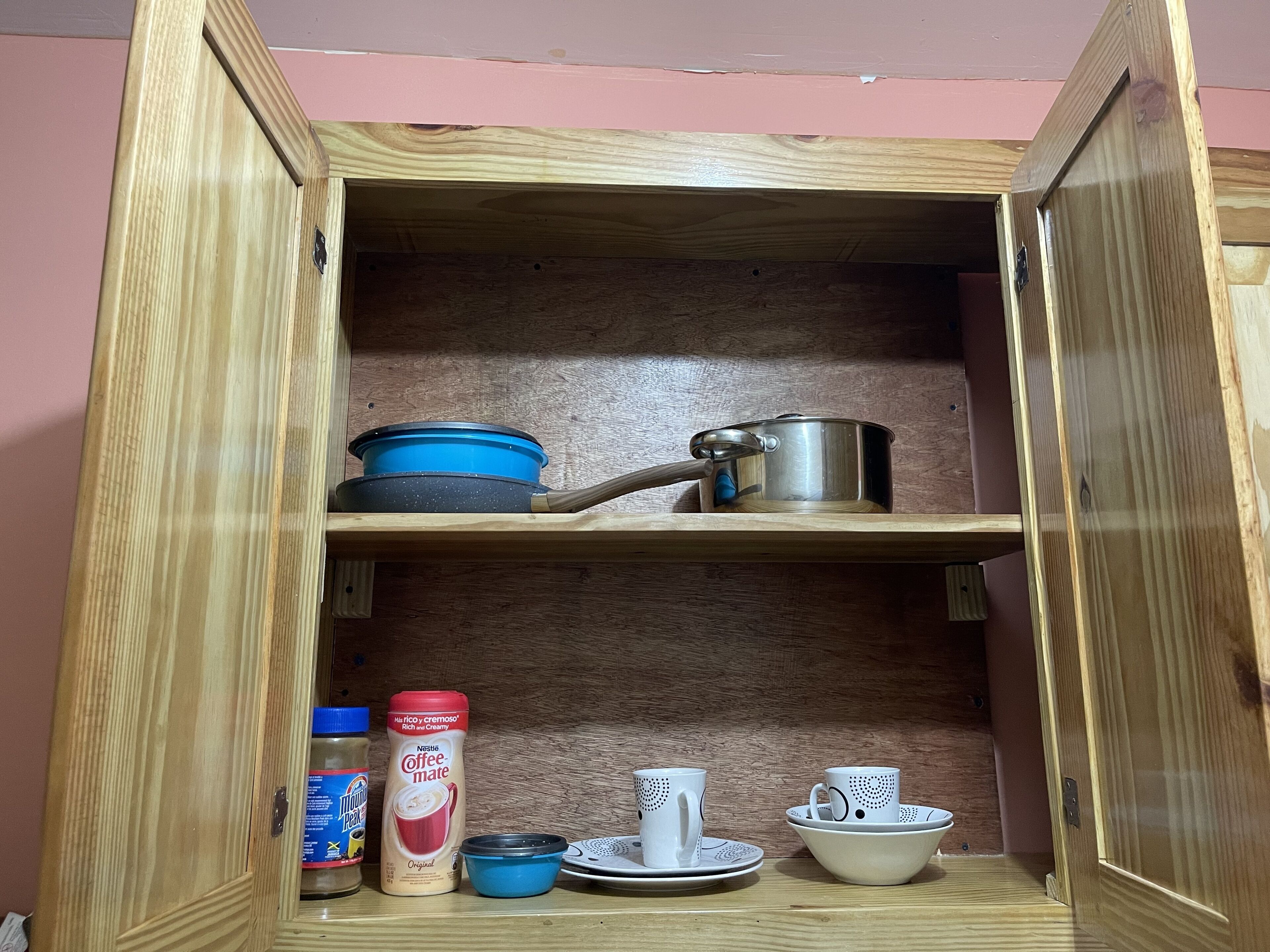 A wooden cabinet with shelves containing kitchenware, including a blue pot, a stainless steel pot, a coffee maker, and a jar of coffee creamer.
