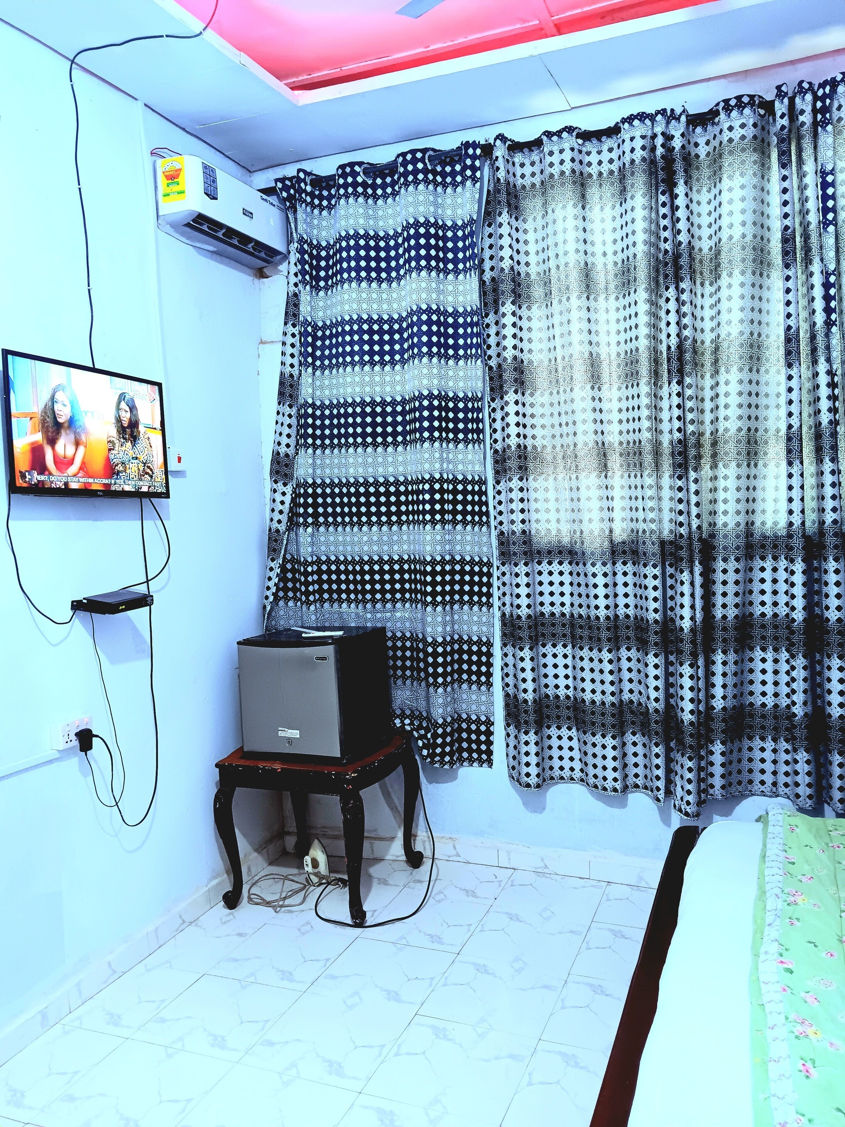 Una habitació amb televisió penjada a la paret, una taula petita amb un ordinador i cortines amb dibuixos.