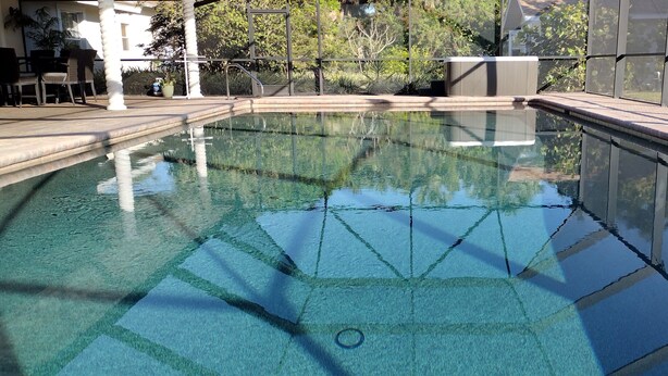 A clear swimming pool with a tiled deck, surrounded by a covered area with chairs and tables.