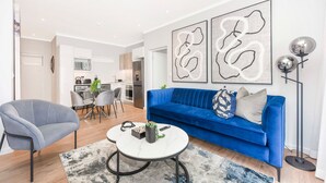 A modern living room with a blue sofa, a grey armchair, and a coffee table. There are abstract wall art pieces and a floor lamp. - Spacious | Brand New | 2 Bed | Rosebank (Johannesburg)